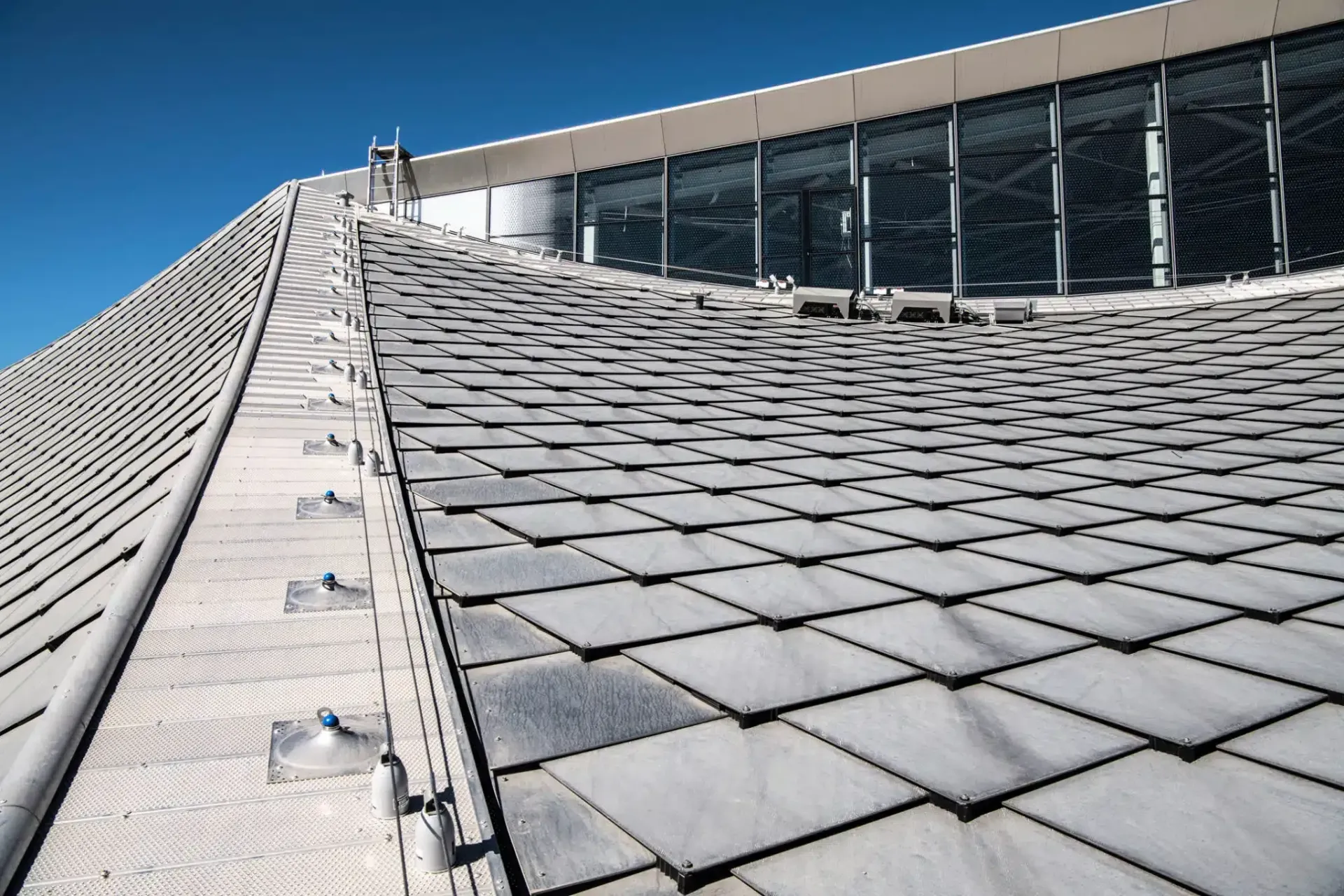 Close-up of a building roof with overlapping gray tiles and a glass-paneled section against a bright blue sky.