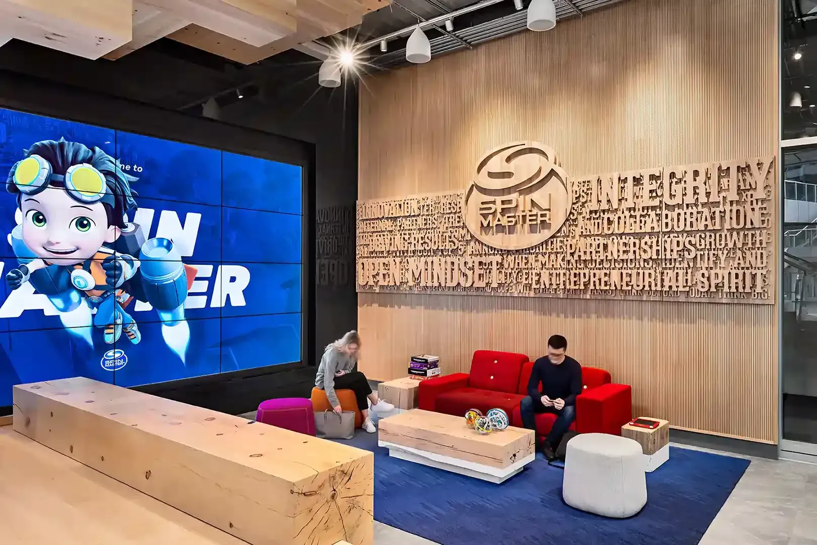 A brightly lit office space with animated character on screen. Two people sit on red sofa and blue carpet. Wooden wall decor.