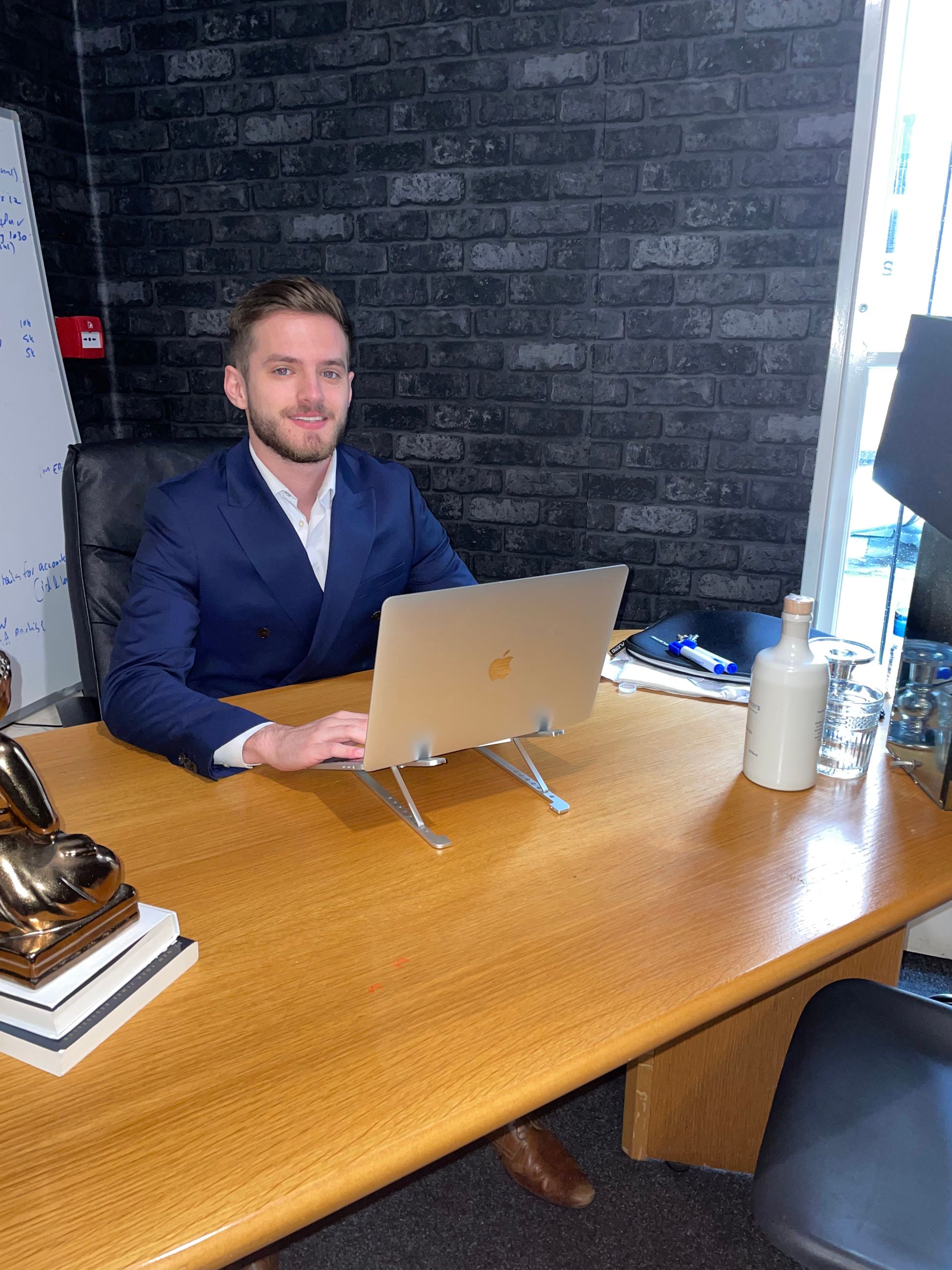 A person in a blue suit sits at a wooden desk with a laptop, smiling in an office with a black brick wall.