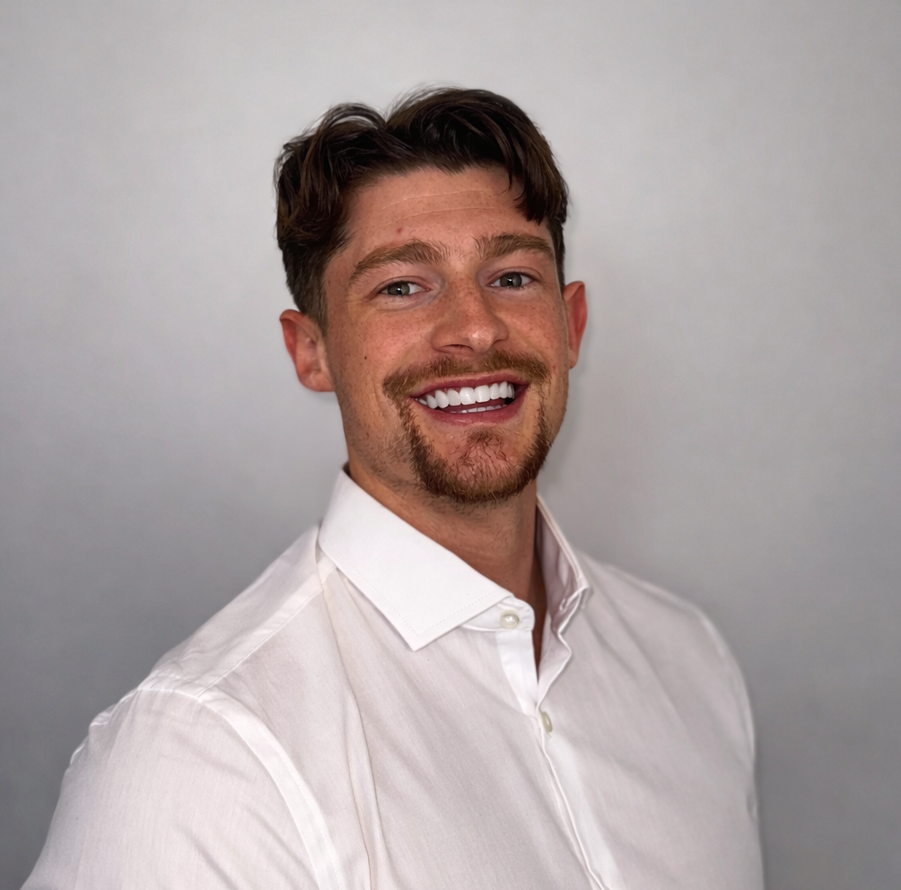 A smiling person with dark hair and a goatee, wearing a crisp white dress shirt against a light gray background.
