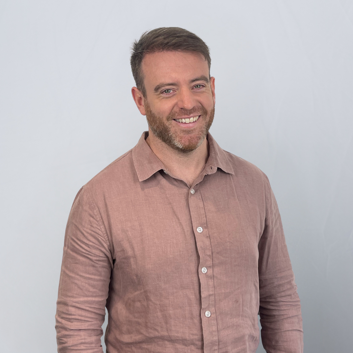 Man with short brown hair and beard, wearing a pink button-up shirt, smiles against a plain white background.