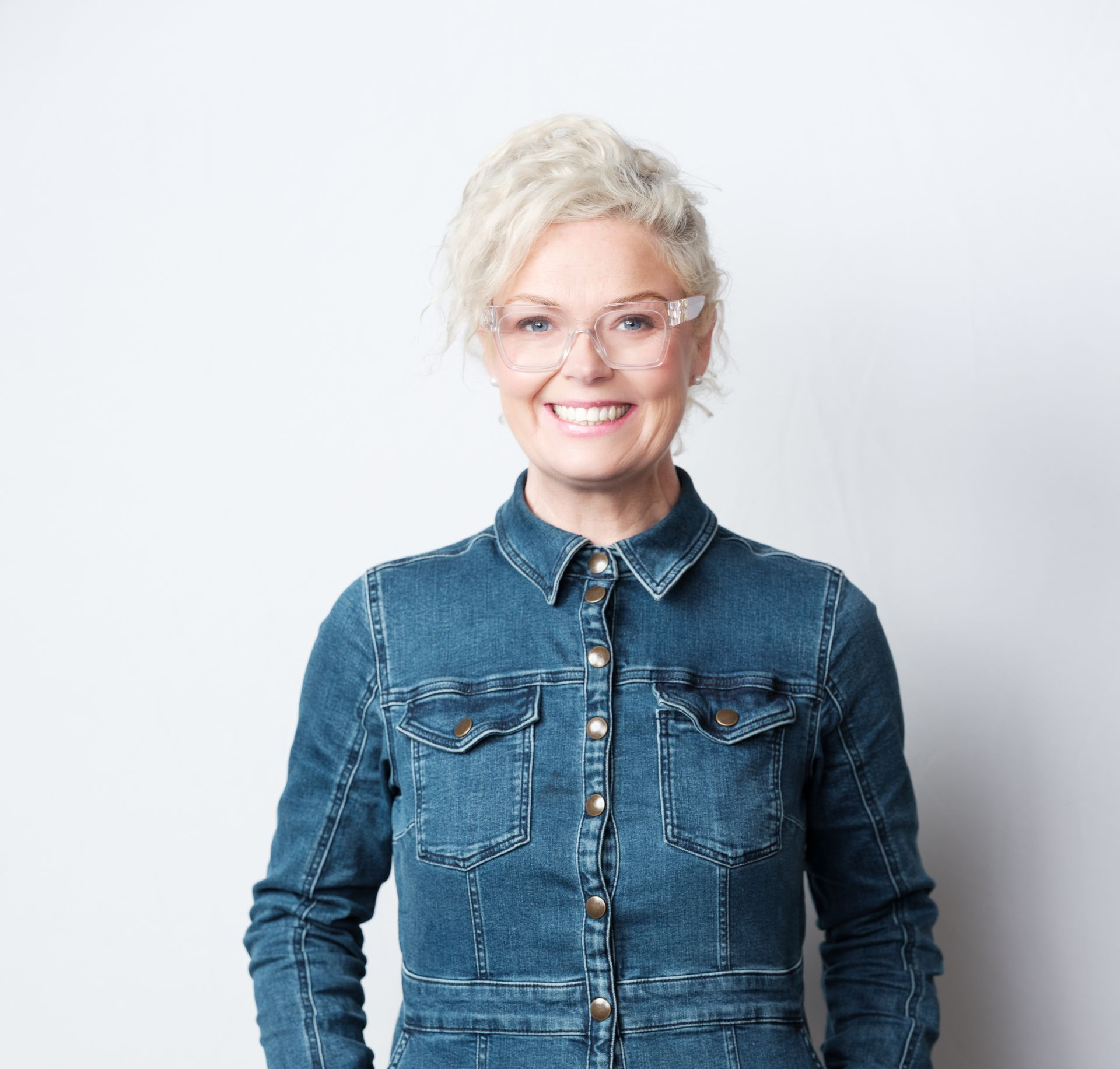 Woman with blonde hair, smiling, wearing glasses and denim jumpsuit. Against a white background.