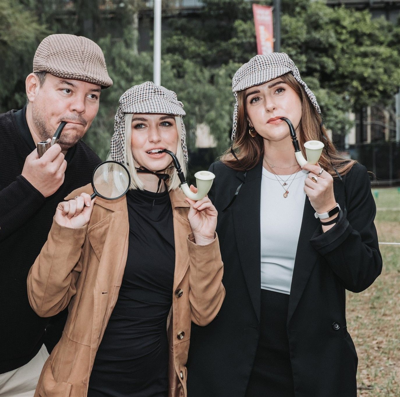 Three people in detective attire holding pipes and a magnifying glass outdoors.