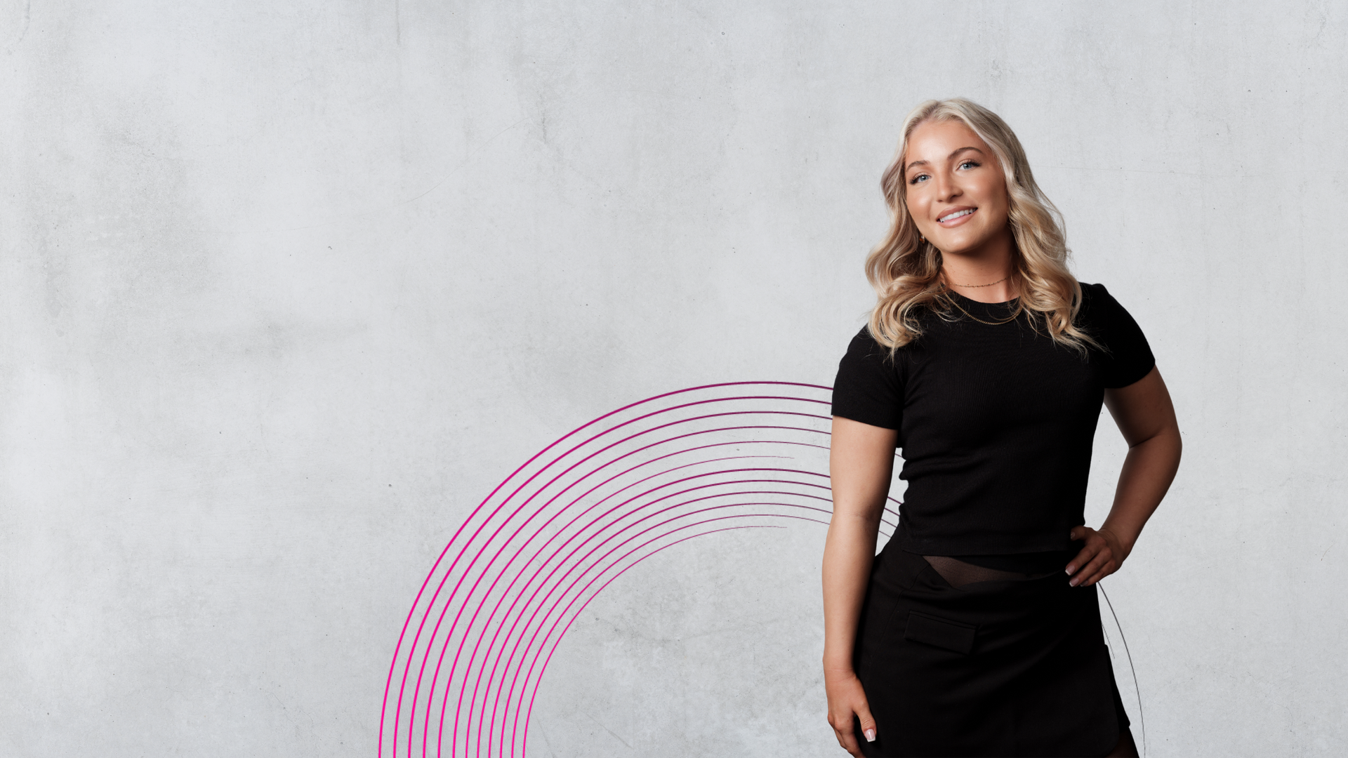 Woman in black attire smiling, hand on hip. Against a white and pink radial background.