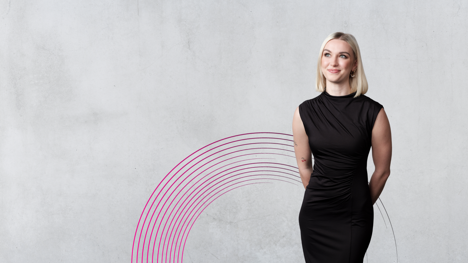 Woman in black dress, smiling, looking upwards, standing against a grey wall with a pink semi-circle design.