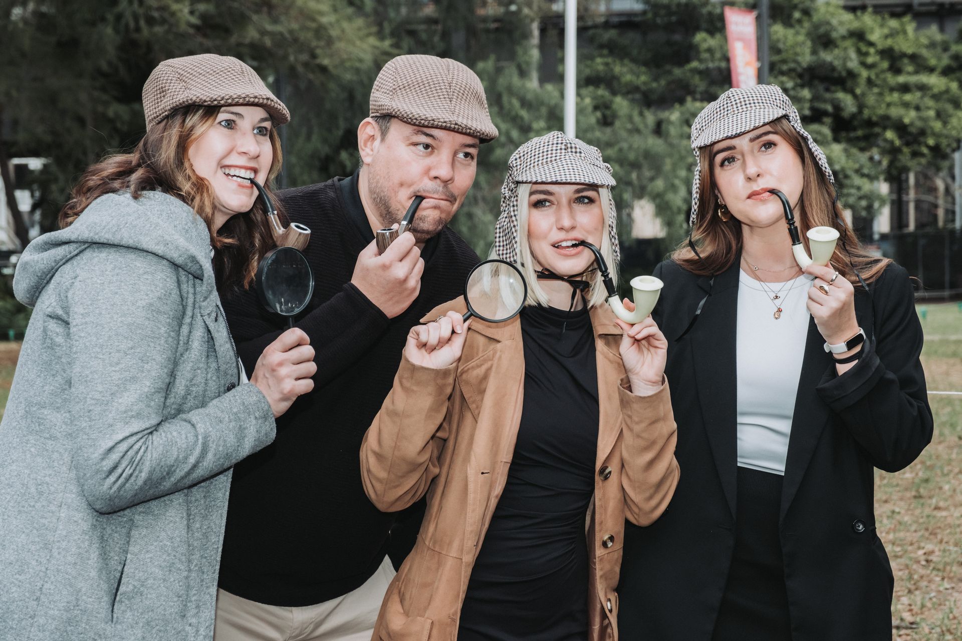 Four people in Sherlock Holmes costumes, posing outdoors with pipes and magnifying glasses.