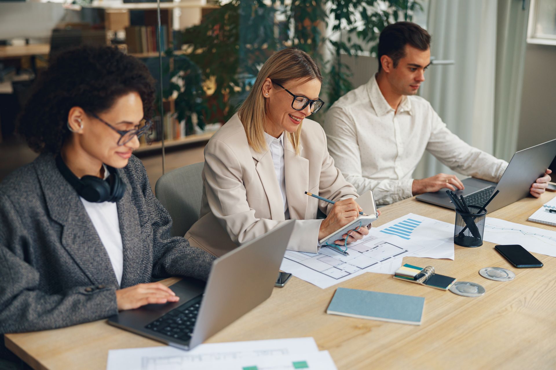 Three coworkers collaborating at a desk with laptops, documents, and a tablet in a bright office