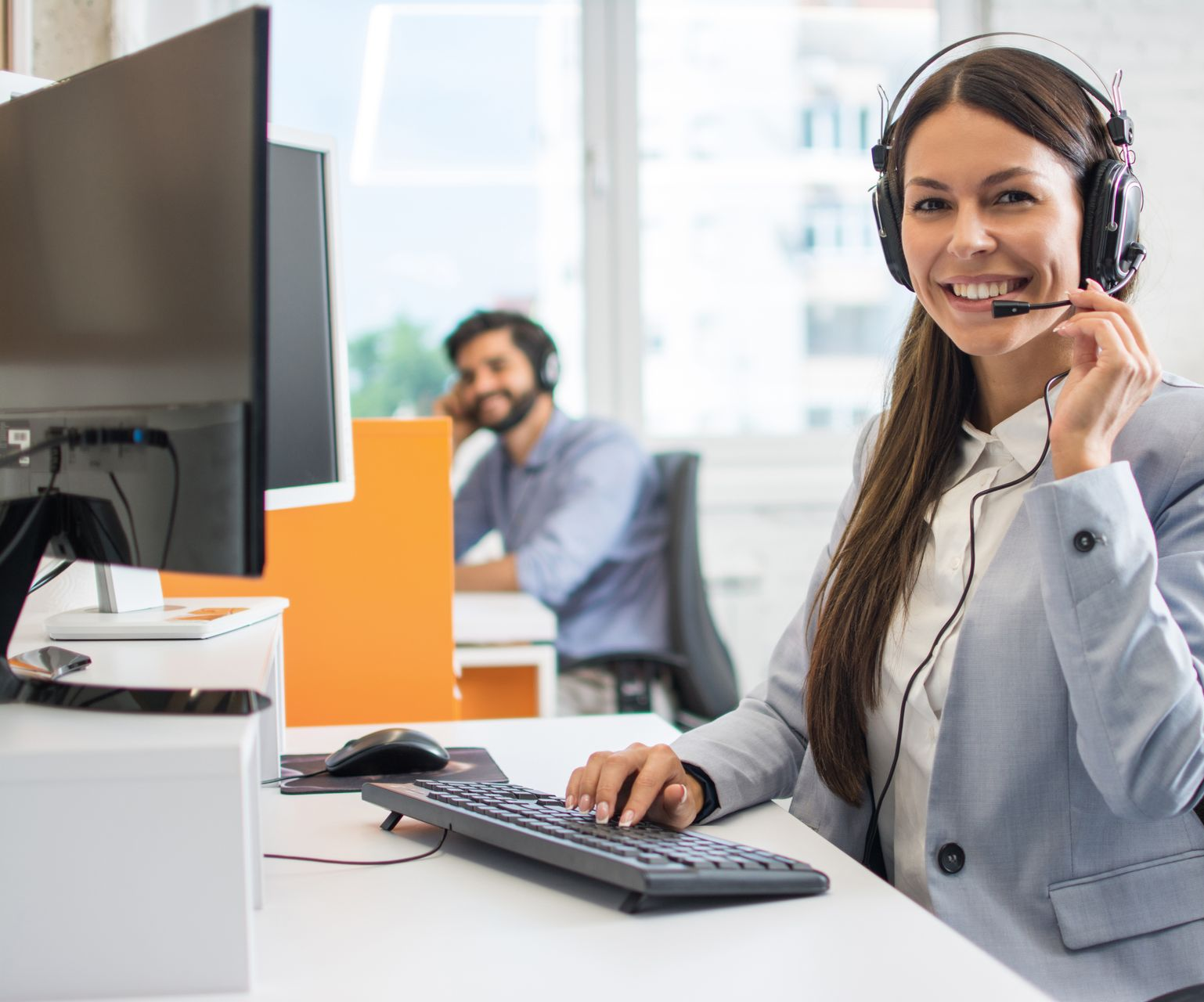 Woman wearing headset at desk, smiling at camera; coworker in background.