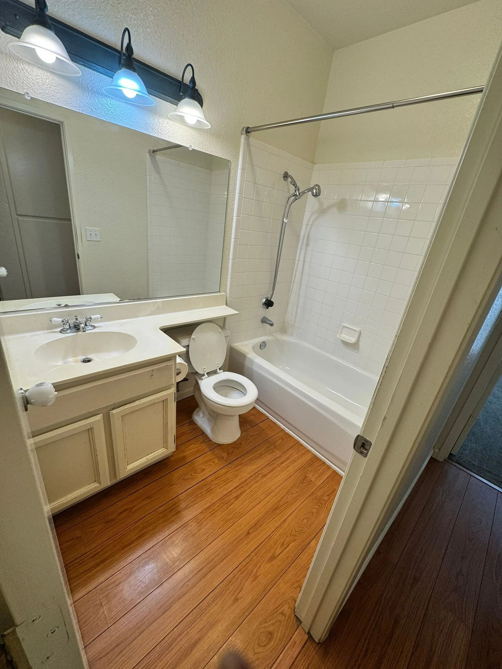 Small bathroom with vanity, mirror, toilet, and shower-tub combo on wood floor.