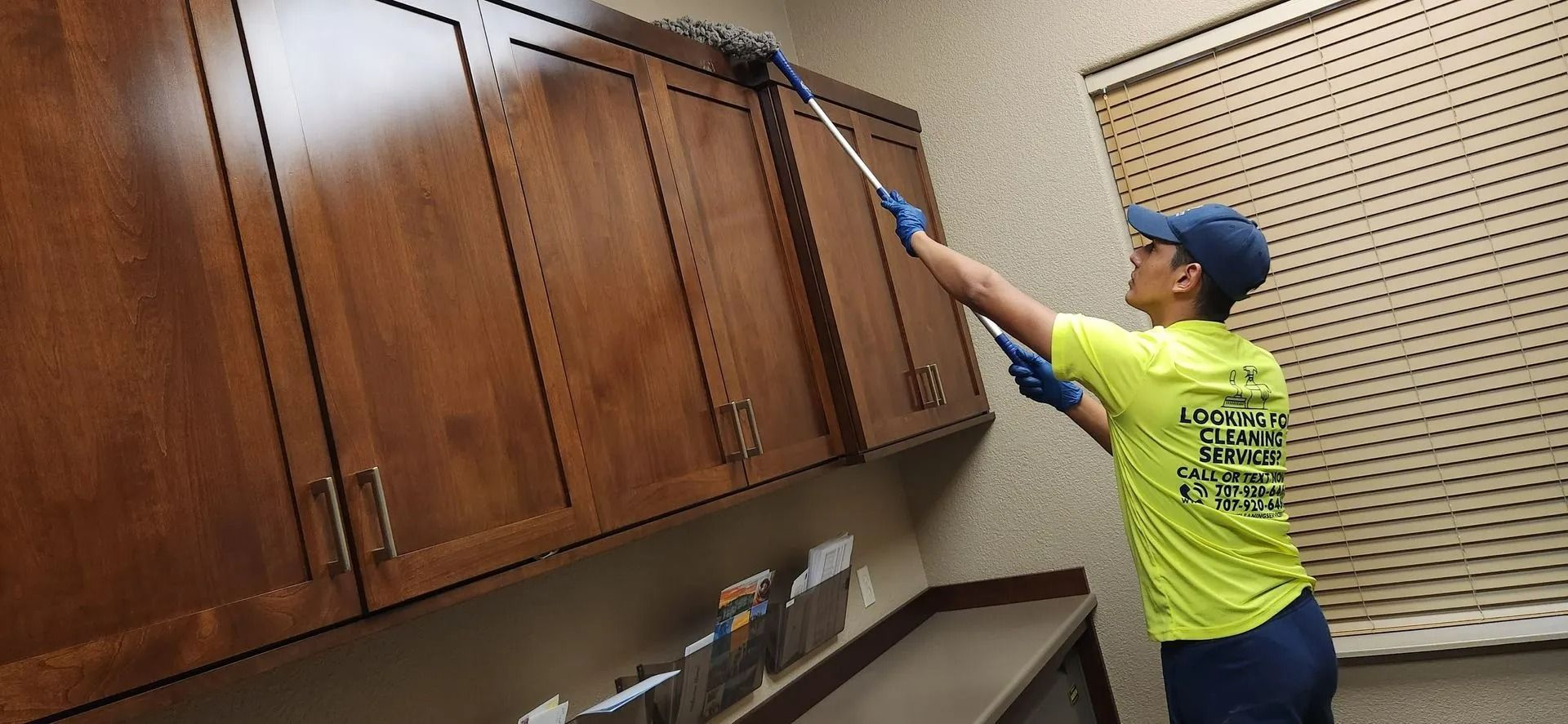 Worker in yellow shirt using a long tool to reach the top of wood cabinets in a store aisle