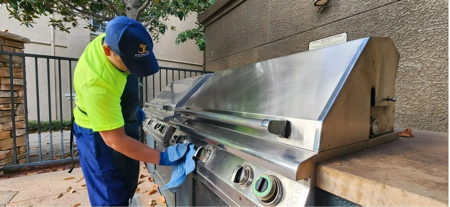 Worker in blue and neon shirt cleaning a stainless steel outdoor grill with a cloth.