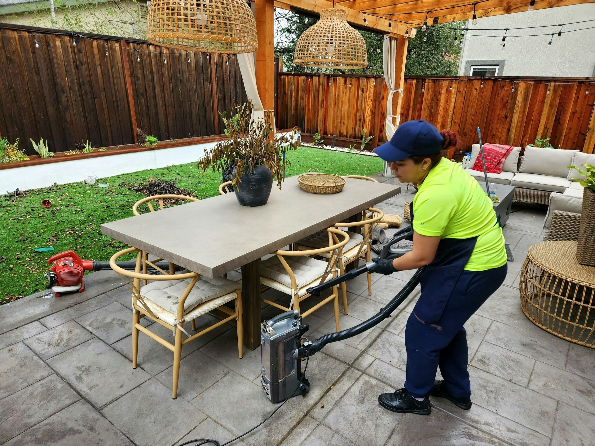 Person pressure-washes a gray patio beside outdoor furniture in a fenced backyard.