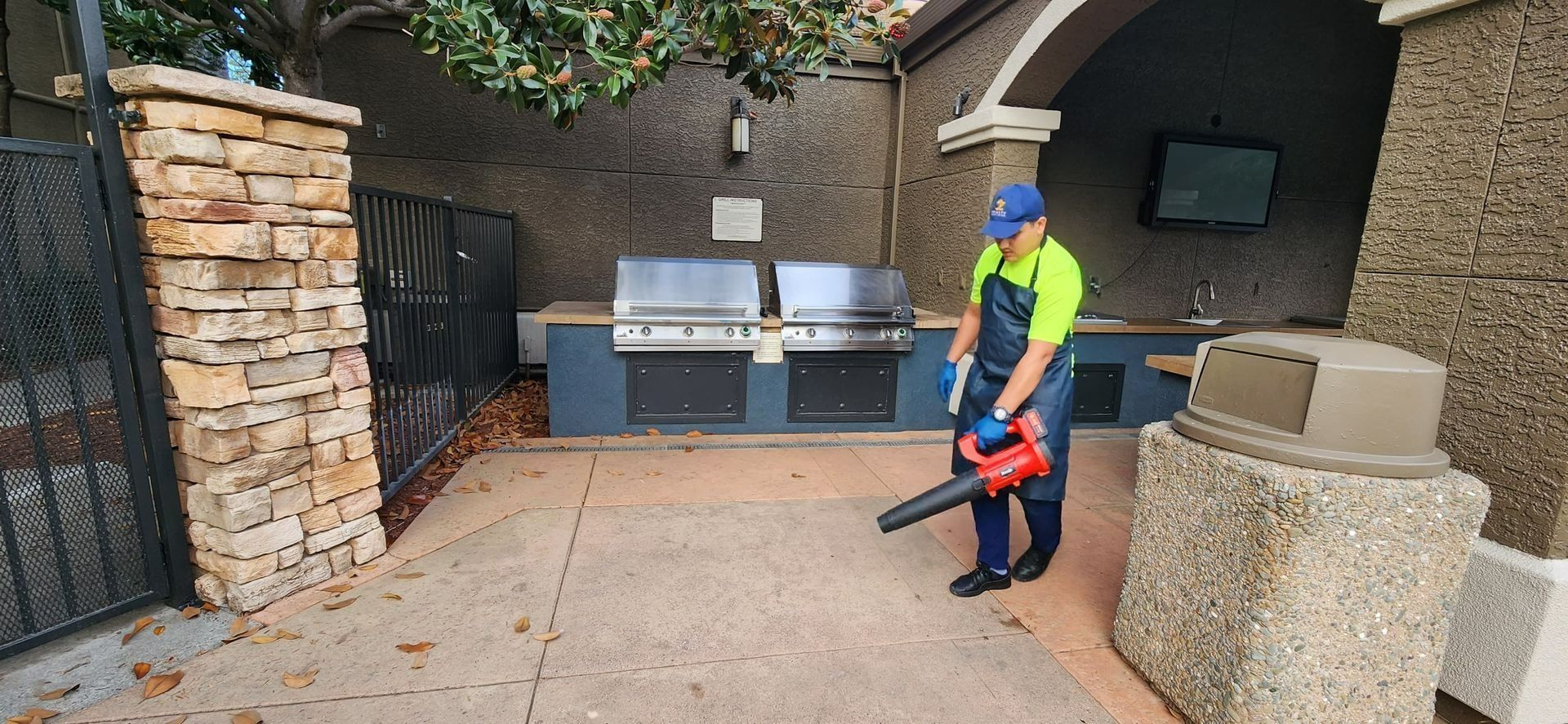 Person using a leaf blower in a stone-paved patio near an outdoor grill and fireplace.