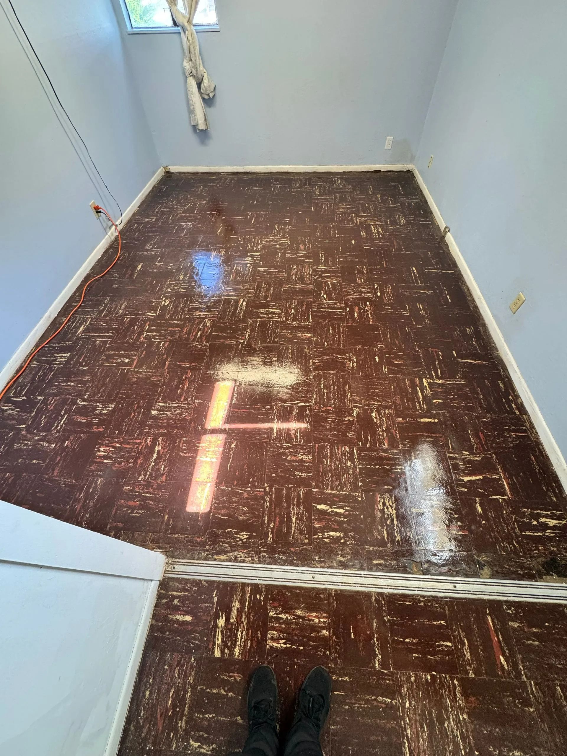 Narrow empty room with brown floor, light gray walls, and a person’s shoes visible at the bottom.