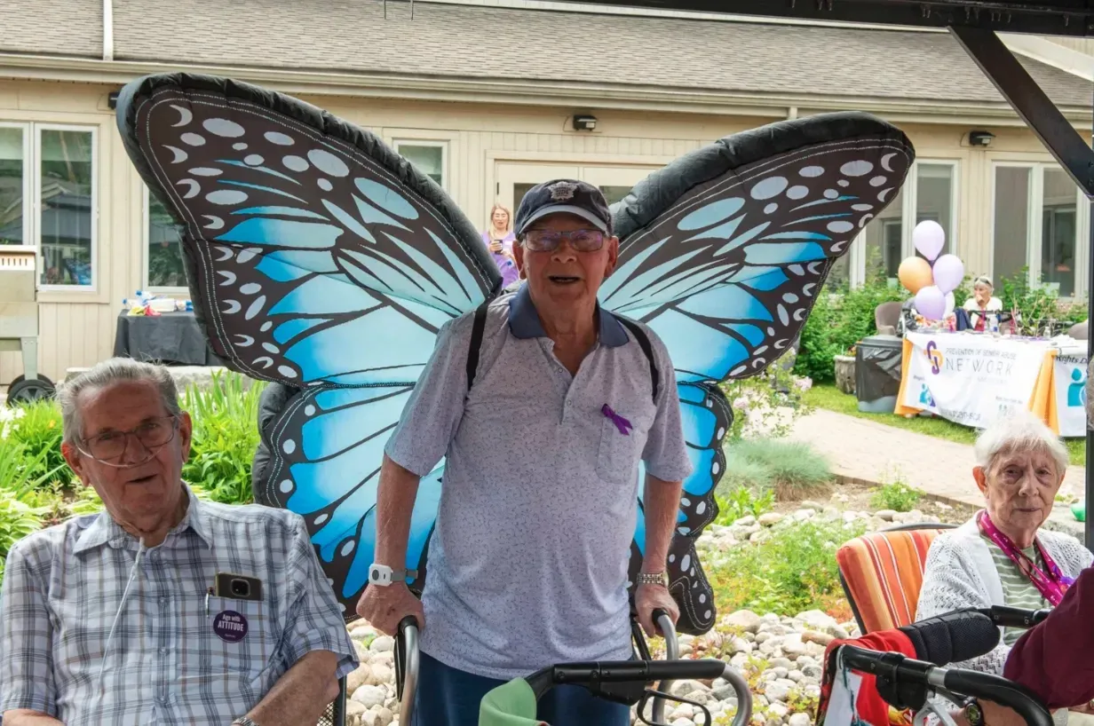 Man with butterfly wings stands between two seated people in a backyard gathering