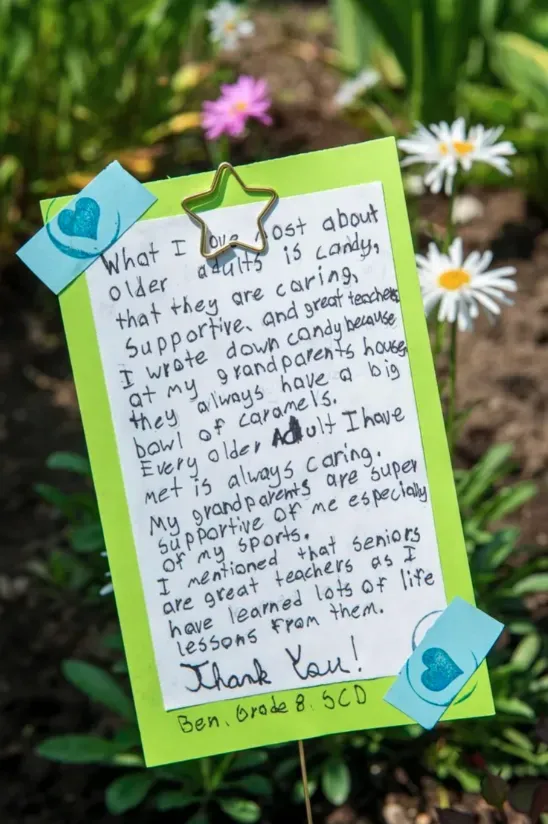 Handwritten thank-you note on green paper clipped outdoors with blue and gold clothespins, flowers blurred behind.