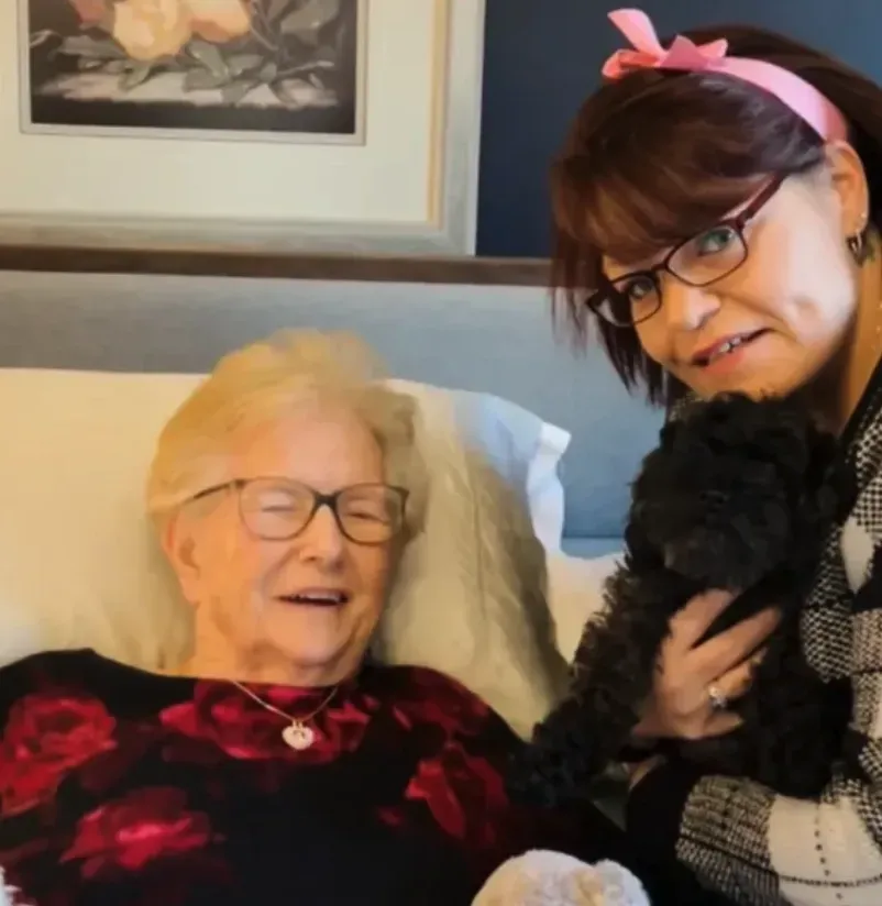 Woman with pink headband holding a small black dog beside an elderly woman smiling in bed