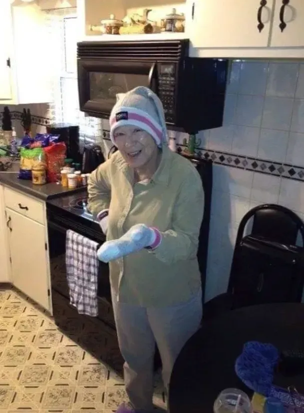 Person in a kitchen wearing a towel hat and gloves, smiling and holding a red object.