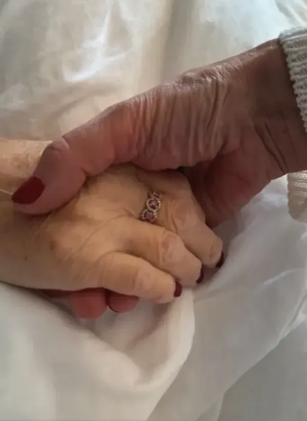Two hands gently holding, one with a ring, on white bedding