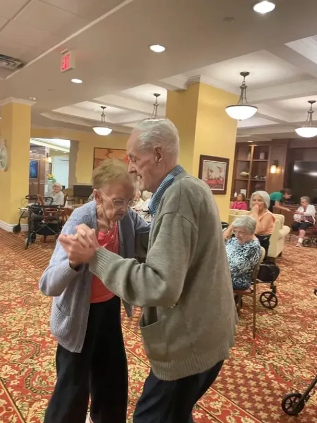 Elderly couple dancing in a warmly lit community hall with seated guests nearby