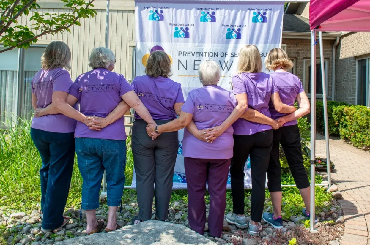 Six people in purple shirts stand with arms around each other at an outdoor event booth.