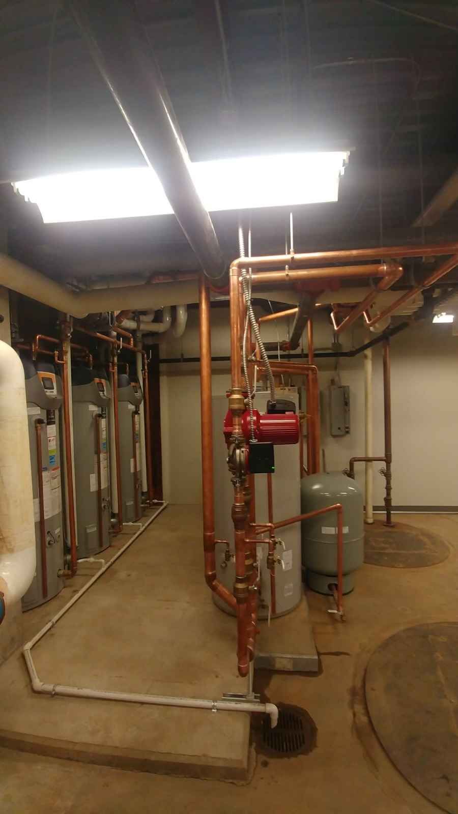 Boiler room with copper pipes, tanks, and a red pump.  Brown floor, white walls, and fluorescent lights.