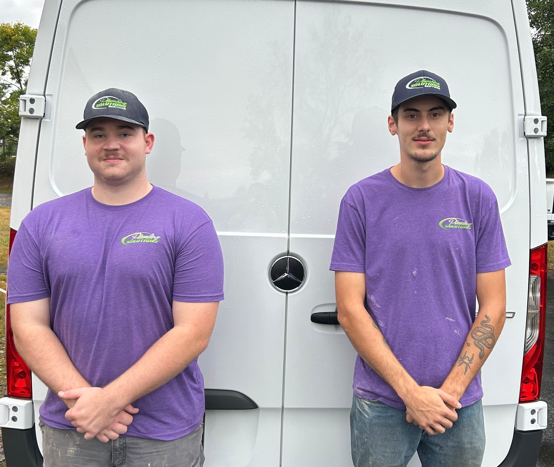 Two men in purple shirts and caps stand in front of a white van.