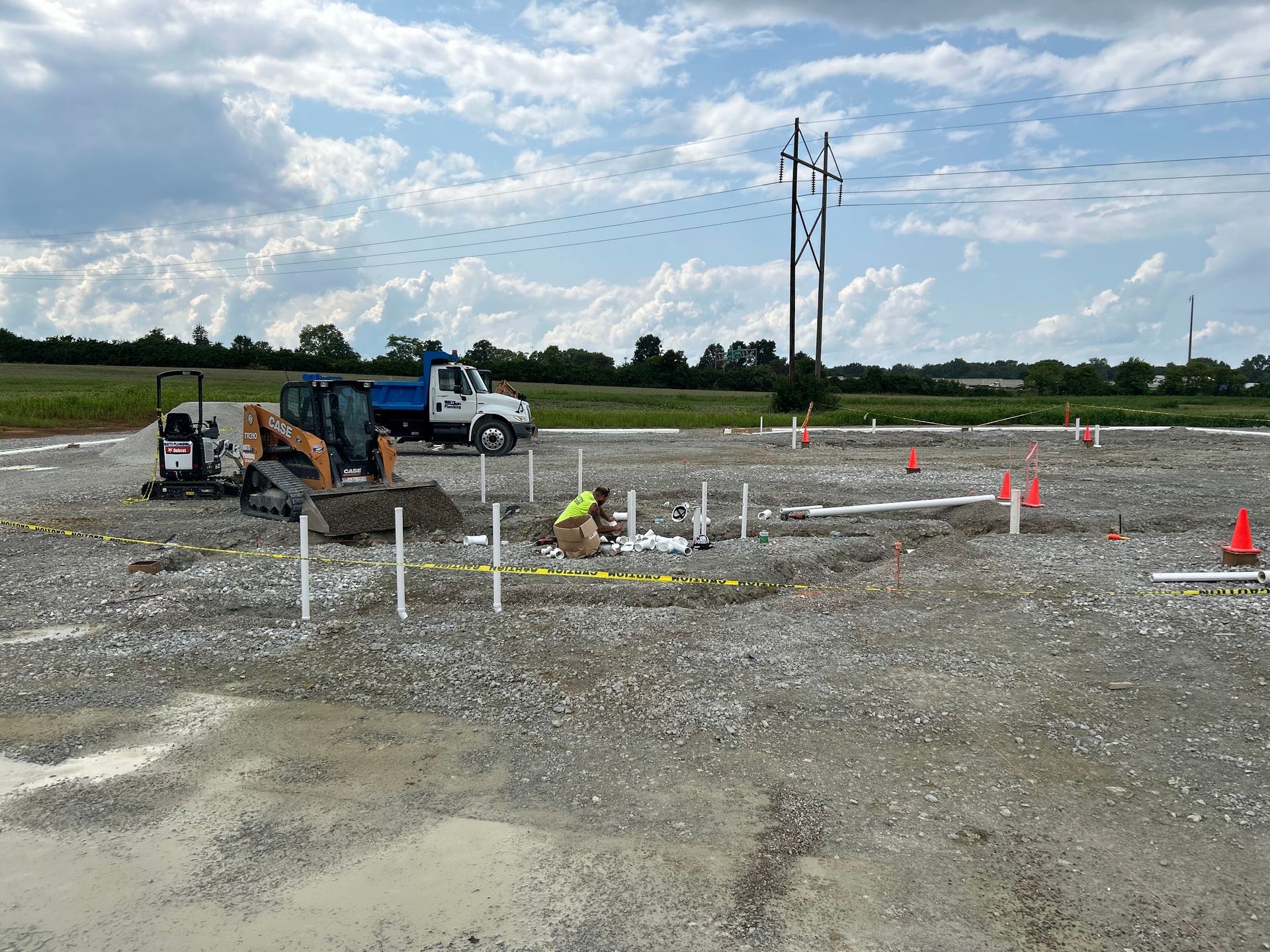 Construction site with pipes, truck, excavator, workers. Cloudy sky, gravel ground.