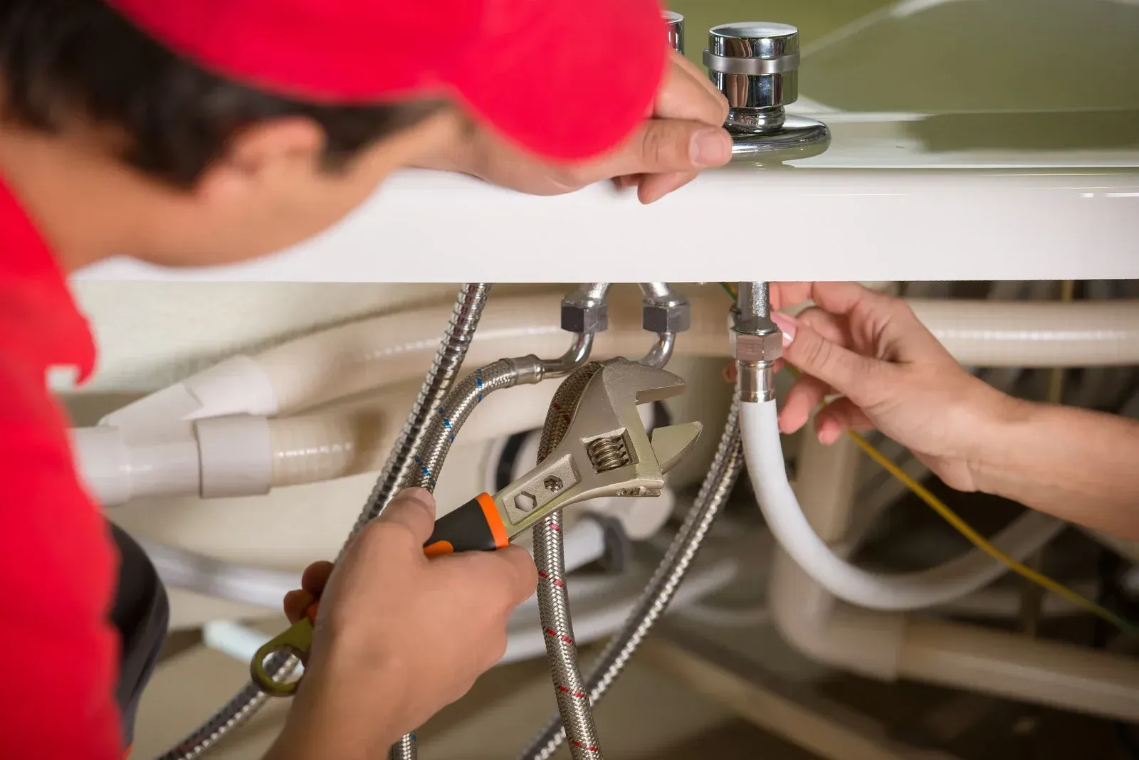 Plumber in red shirt using a wrench to fix pipes under a sink.