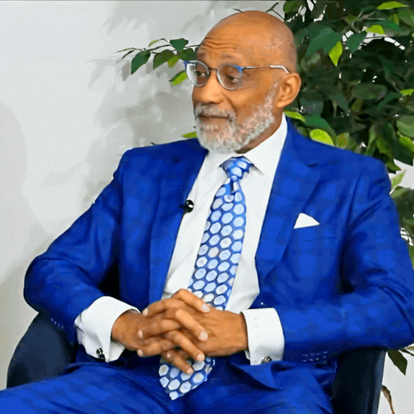 Man in blue suit, glasses, and tie, seated, speaking, hands clasped, indoor setting with plant. Andre Bartell.