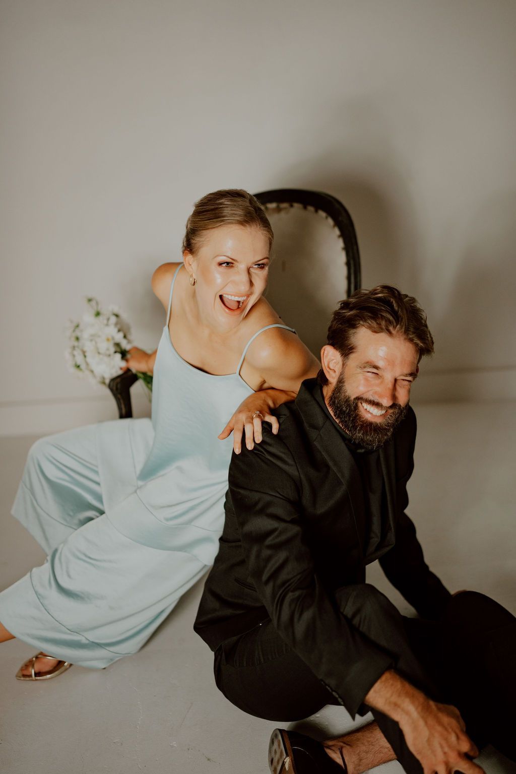 A woman in a light blue jumpsuit and a man in a black suit are laughing together near a chair. They are posing indoors.