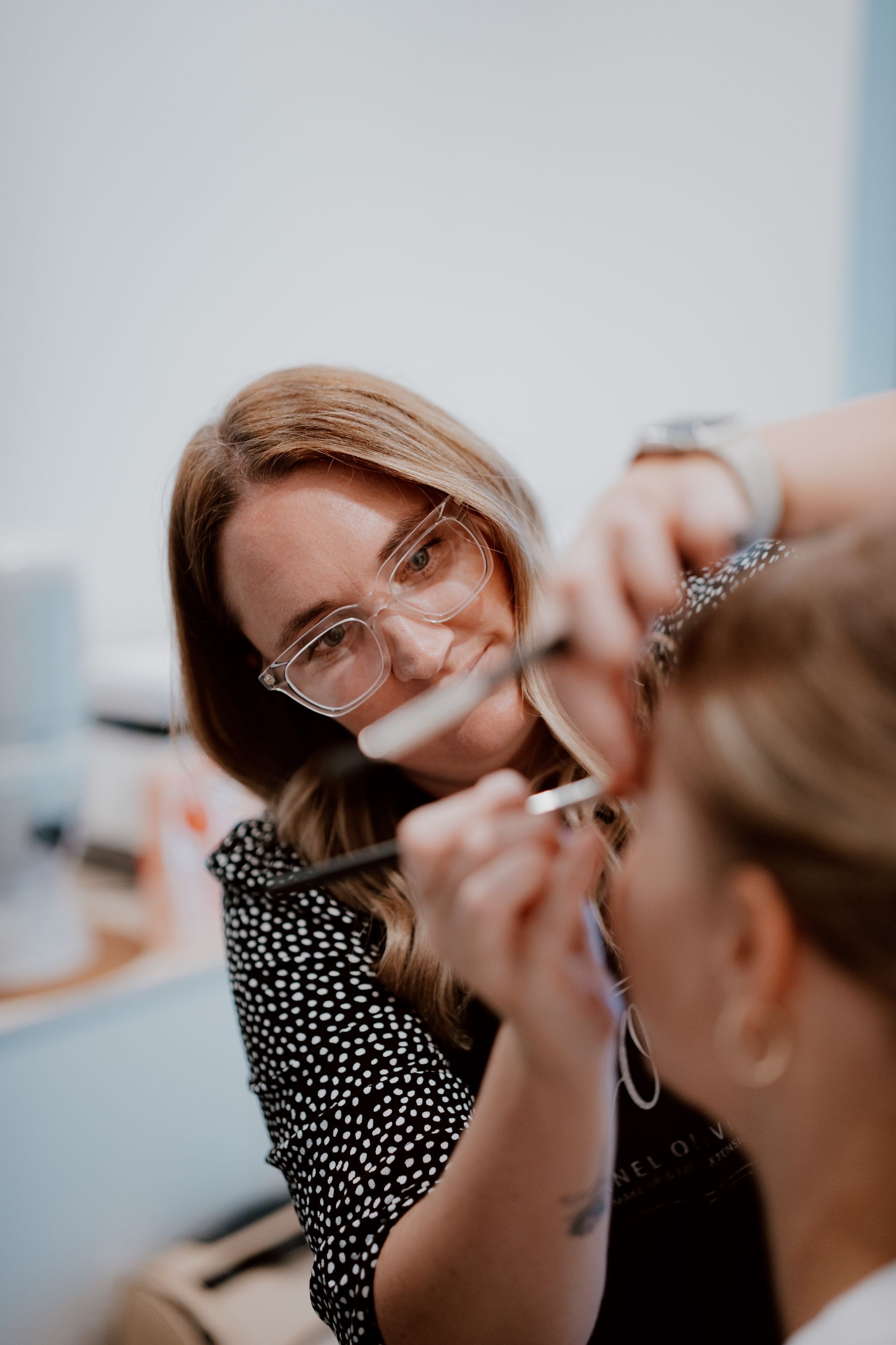 Makeup artist applying eyeshadow with a brush, leaning over a client. She wears glasses and a polka-dot dress.
