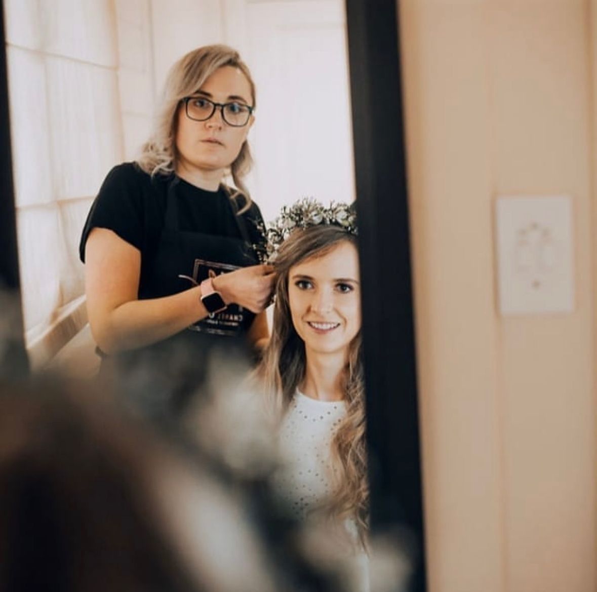 Hair stylist with glasses arranges the bride's hair, both reflected in a mirror. The bride smiles in a white dress.
