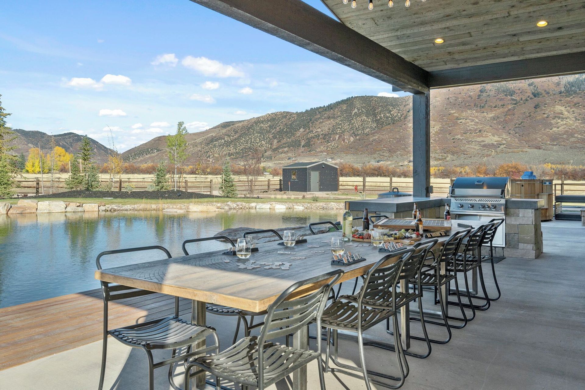 Outdoor patio with long table and grill, overlooking pond and mountains.