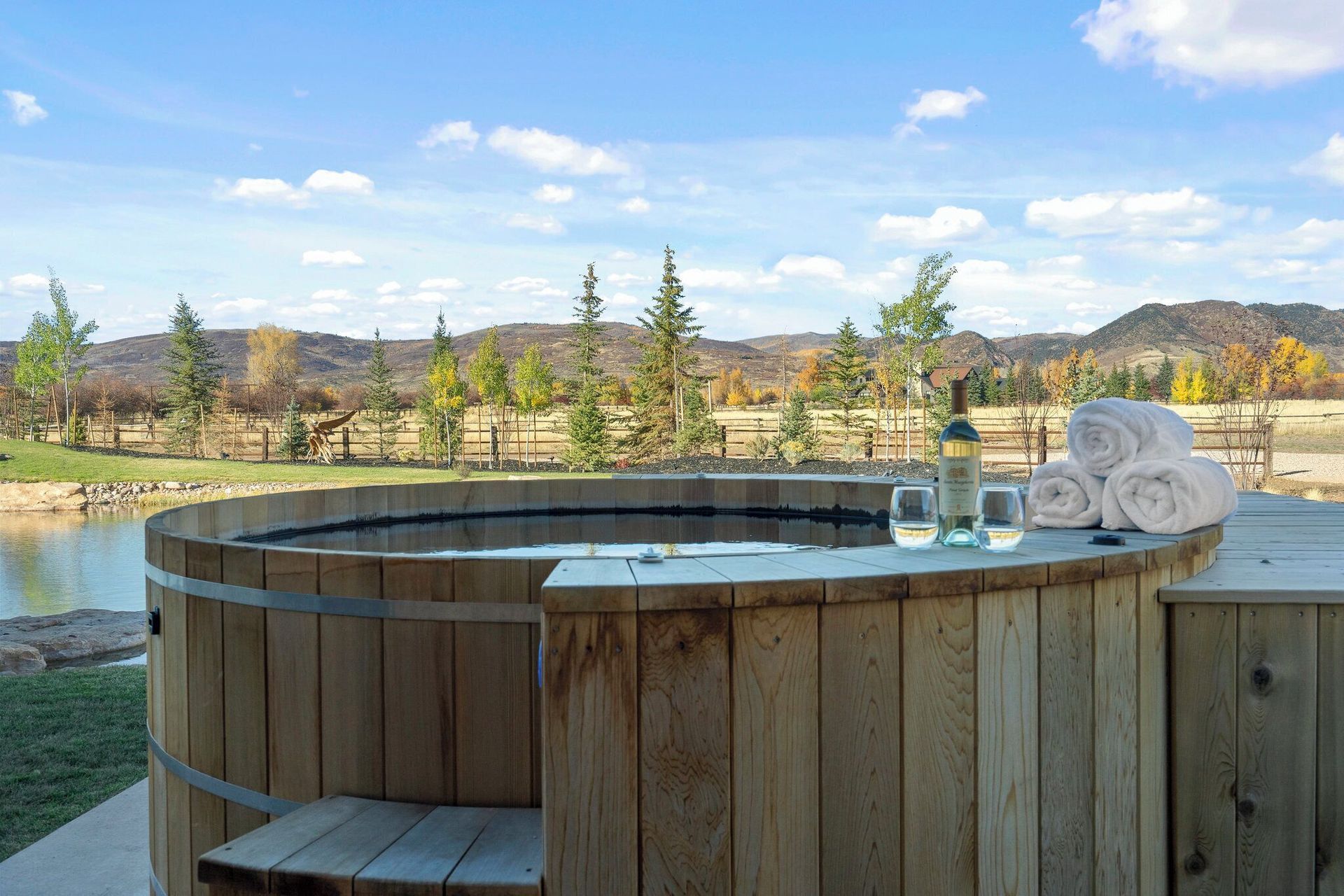 Wooden hot tub outdoors with towels, wine, and a scenic view of trees and water.