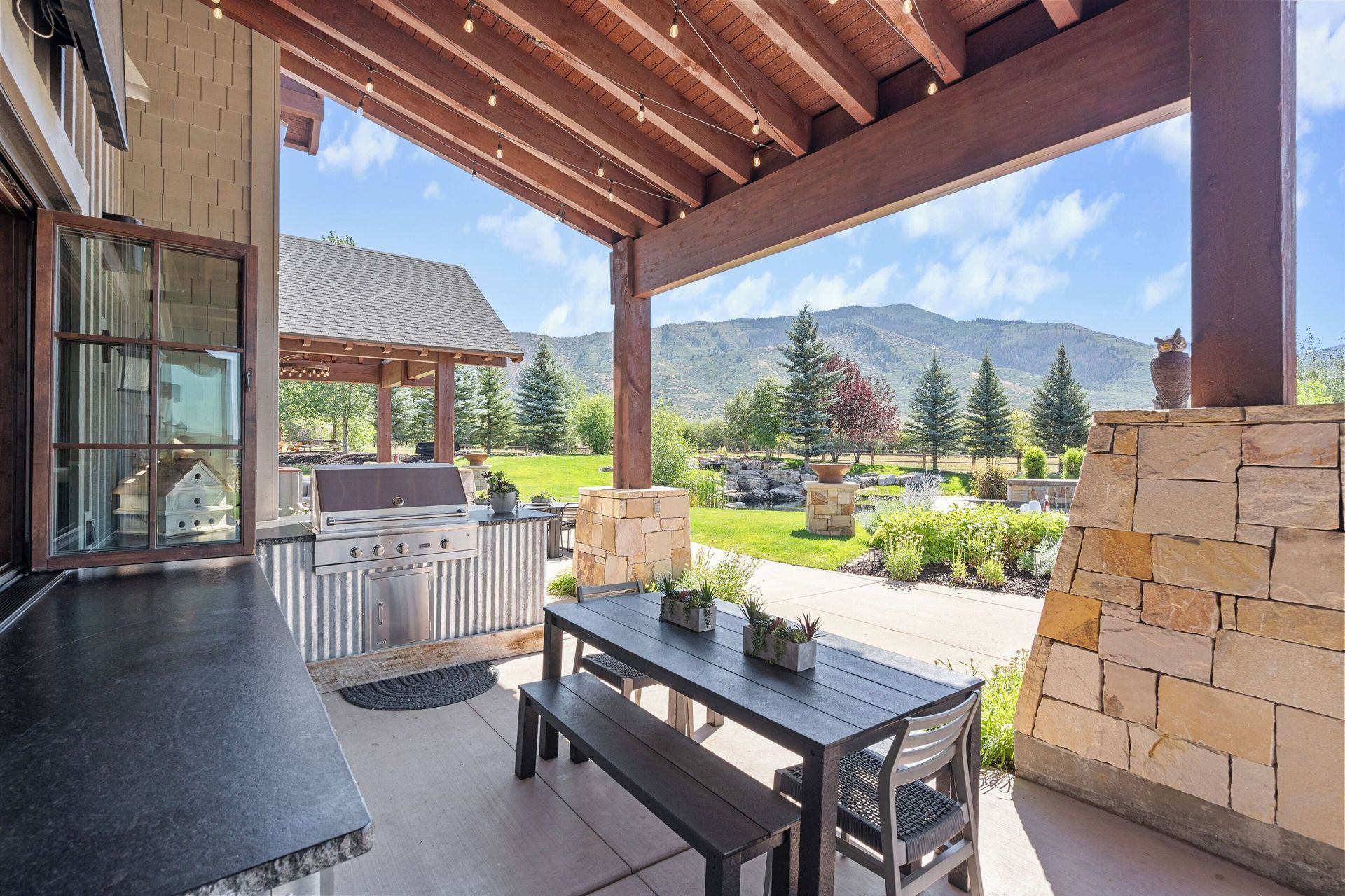Patio with outdoor kitchen, dining table, mountains in the background. Sunny day, modern design.