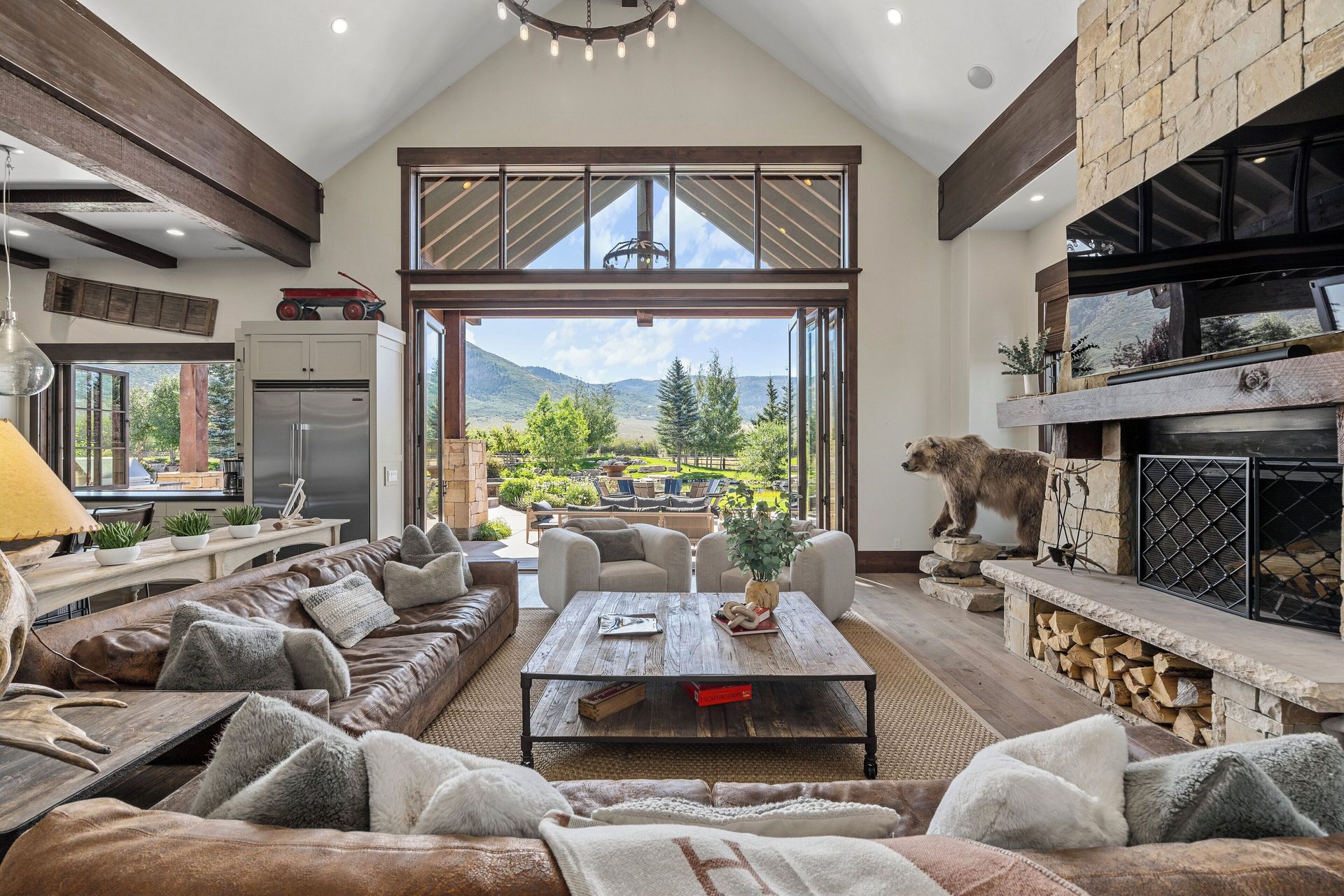 Spacious living room with open glass doors, mountain view. Leather sofa, stone fireplace, and bear statue.