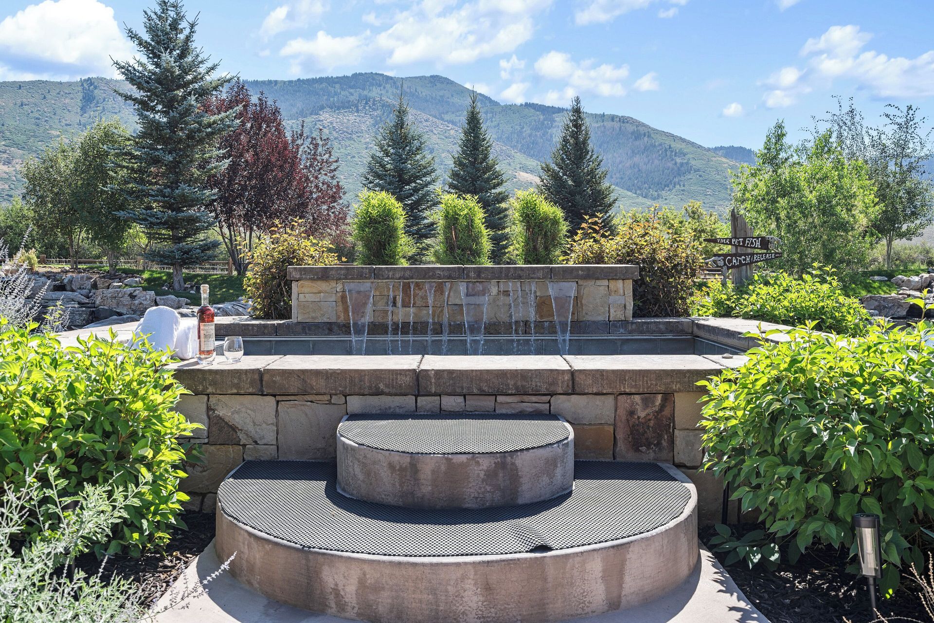 Stone fountain in a garden setting with mountain backdrop and blue sky.