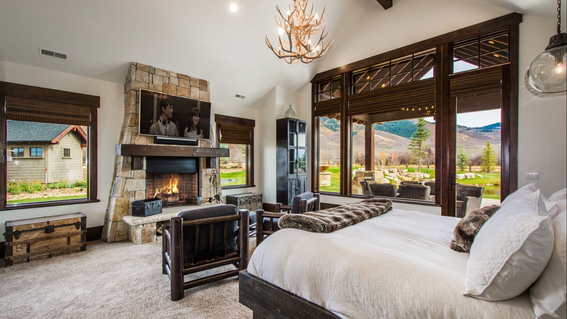 Bedroom with fireplace, mountain view, rustic decor: stone, wood, neutral colors, large bed, windows, chandelier.