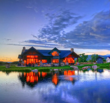 House with orange interior lights reflecting on lake at sunset, under cloudy sky.