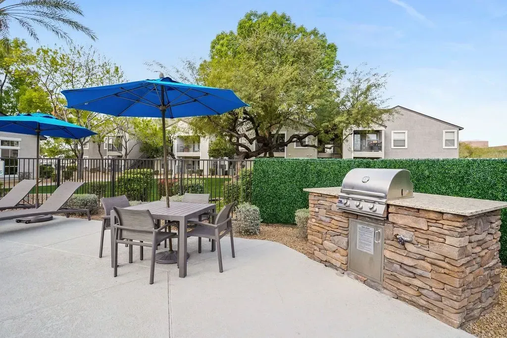 Outdoor grilling station with a stone base and stainless steel grill, adjacent to a table and chairs under blue umbrellas, with apartment buildings in the background at Heritage at Deer Valley in Phoenix, AZ.
