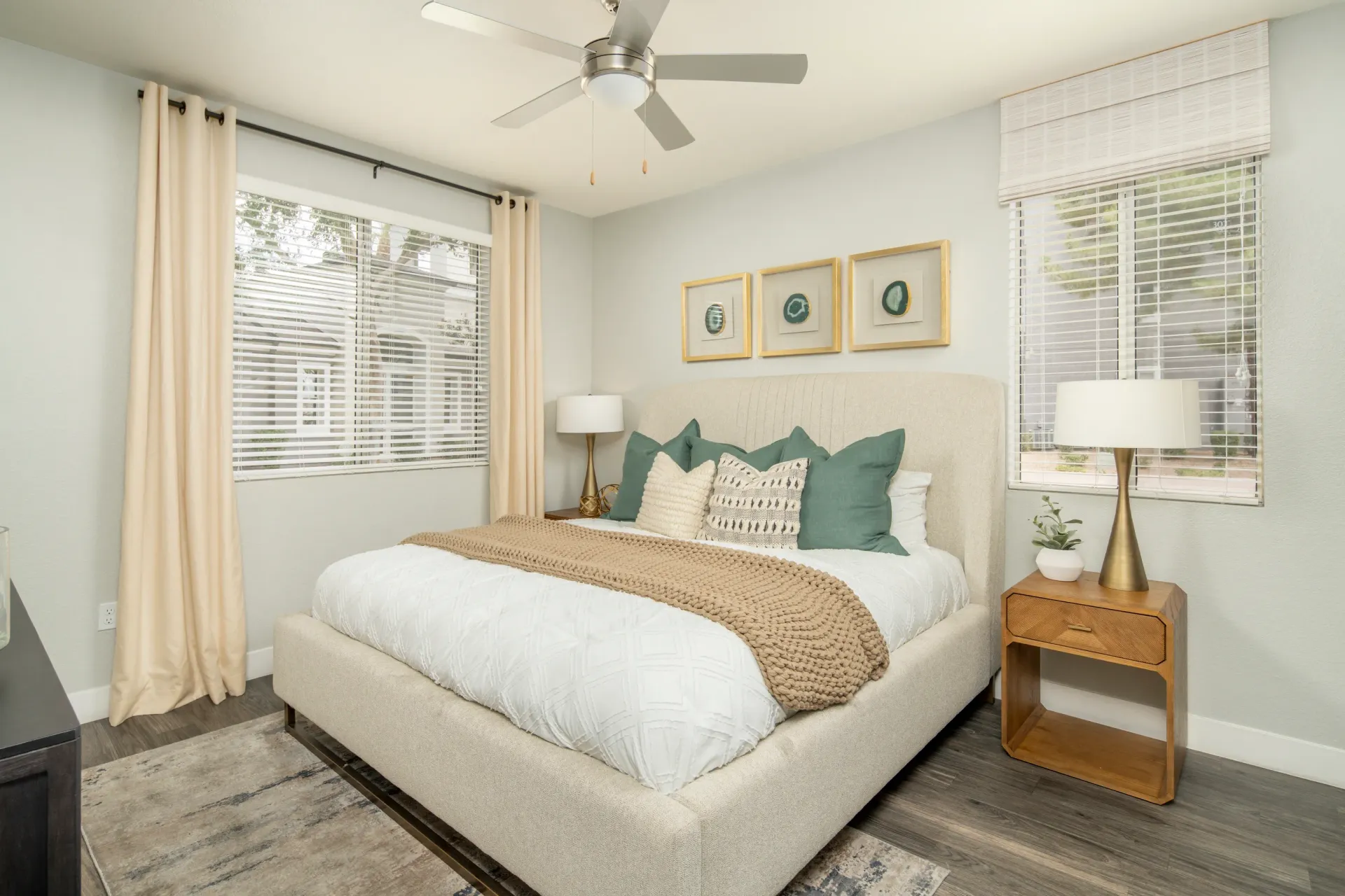 A king-sized bed with white bedding and a tan throw, a ceiling fan, and artwork in a bedroom at Heritage at Deer Valley in Phoenix, AZ.