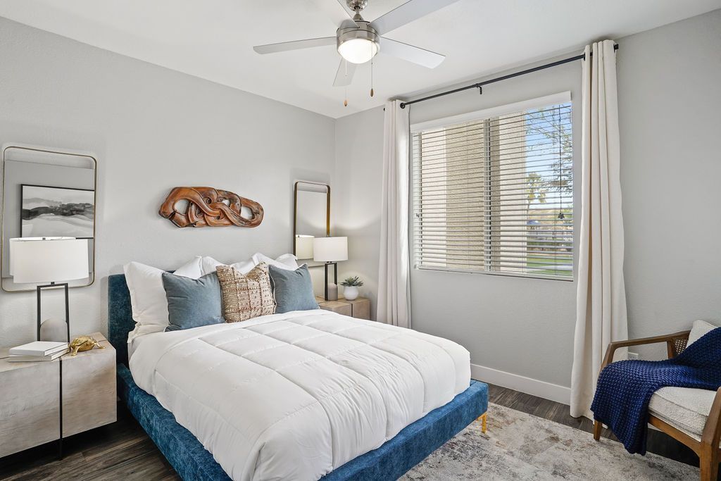 Modern apartment bedroom with a blue upholstered bed, white bedding, two nightstands, and a window with blinds at Heritage at Deer Valley in Phoenix, AZ.