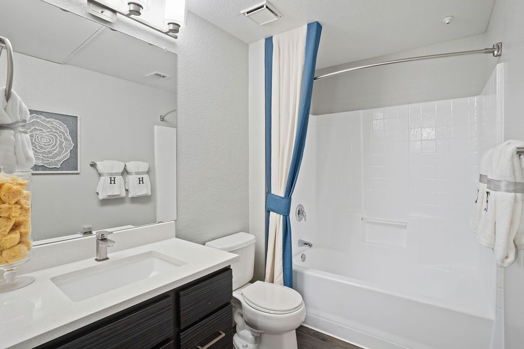 Bathroom with white sink, mirror, toilet, and tub/shower with a blue-and-white curtain at Heritage at Deer Valley in Phoenix, AZ.