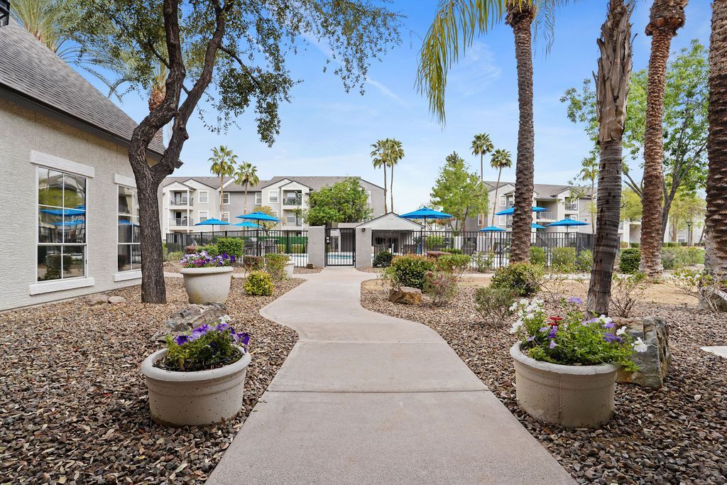 Courtyard of an apartment complex with a paved path, palm trees, planters, and a fenced pool at Heritage at Deer Valley in Phoenix, AZ.