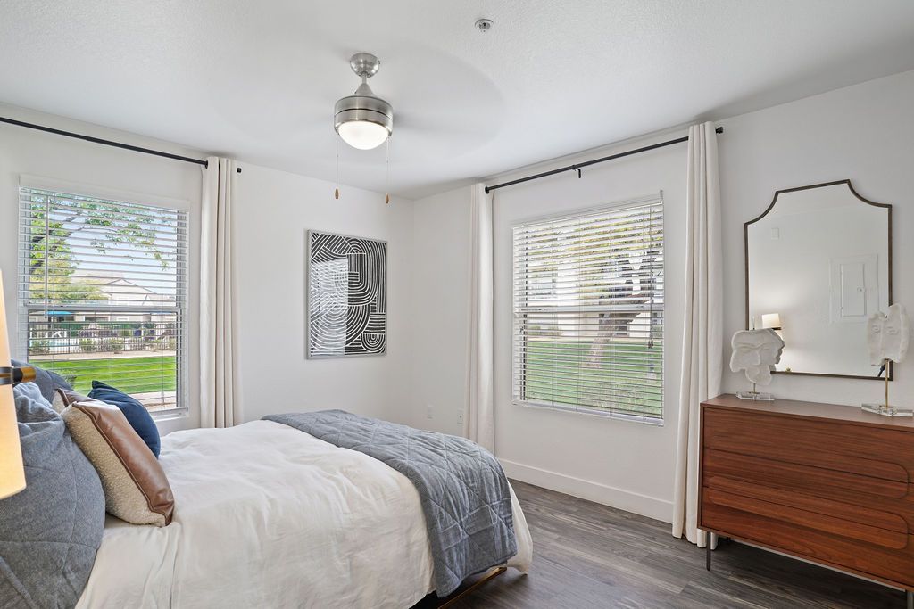 Bright bedroom in a modern apartment with a bed, dresser, mirror, and two windows with blinds at Heritage at Deer Valley in Phoenix, AZ.
