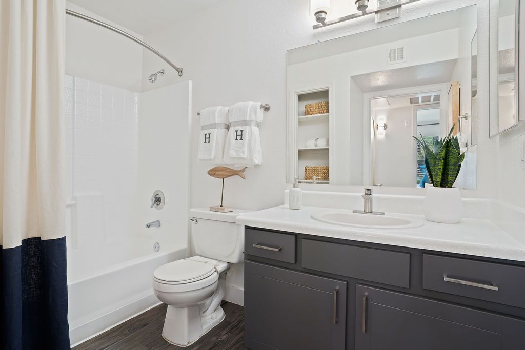 Modern bathroom with white vanity, dark gray cabinets, large mirror, toilet, and tub with shower curtain at Heritage at Deer Valley in Phoenix, AZ.