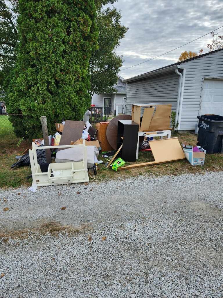 Outdoor clutter of furniture, boxes, and debris piled beside a white house and tree on a gravel driveway