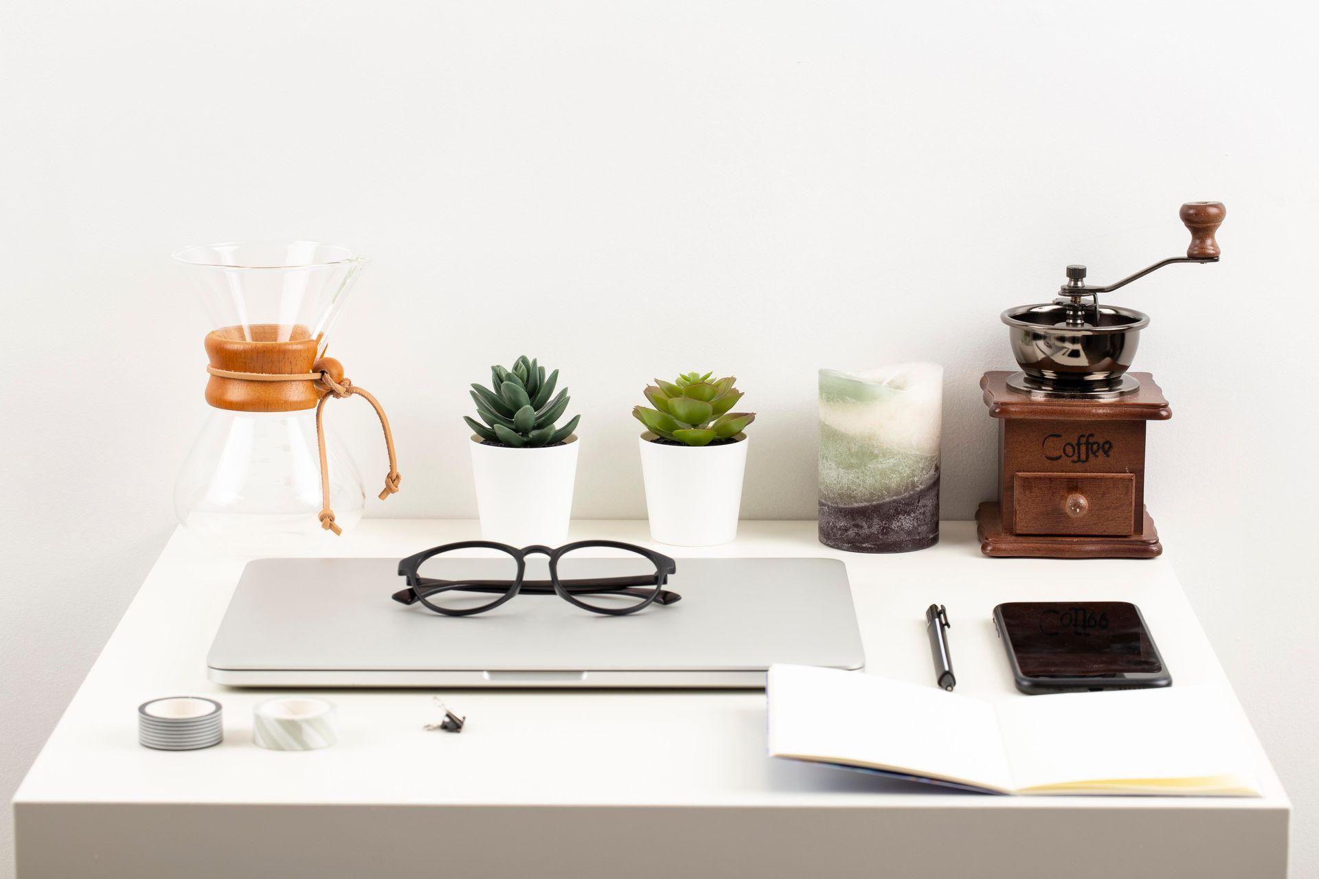 A desk with a laptop , glasses , plants , a coffee grinder and a candle.