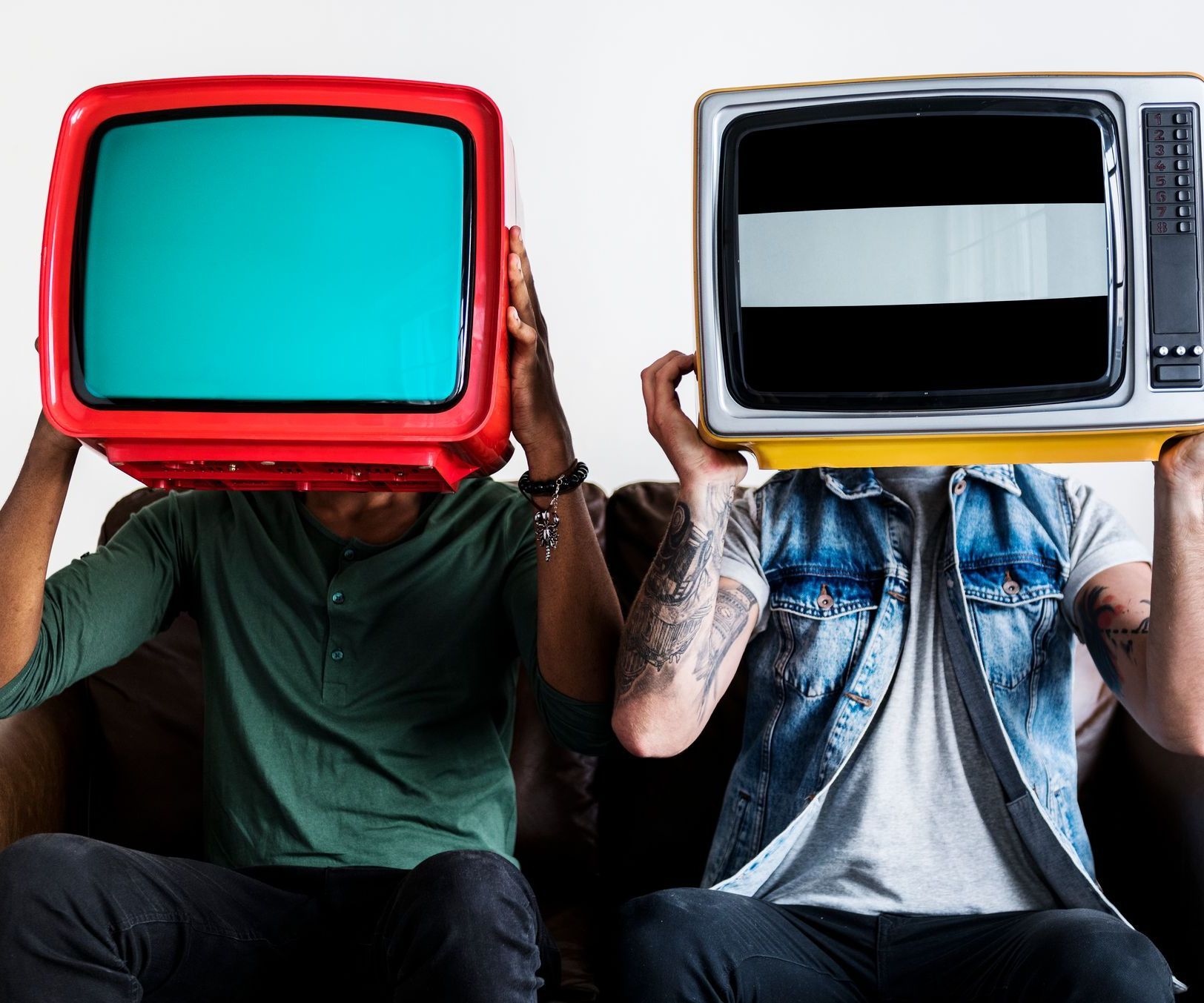 Two men are sitting on a couch holding televisions in front of their faces
